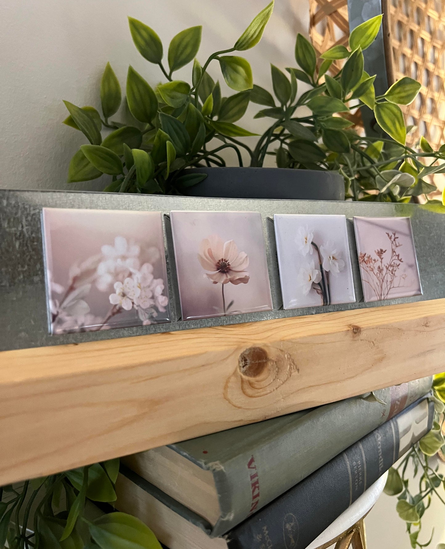 Stack of books with a wooden cover stand featuring small square images of flowers, surrounded by green plants. magnets